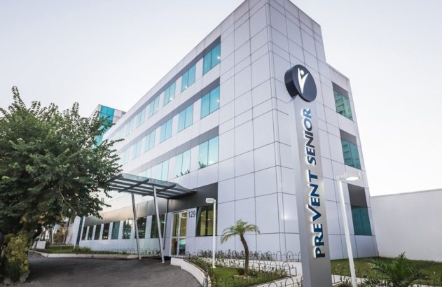 Prédio moderno de fachada clara e janelas de vidro, com placa vertical "PREVENT SENIOR" na frente.