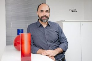 Homem barbudo sorrindo, apoiado em balcão com vasos em escritório. Logo 'invest news'.