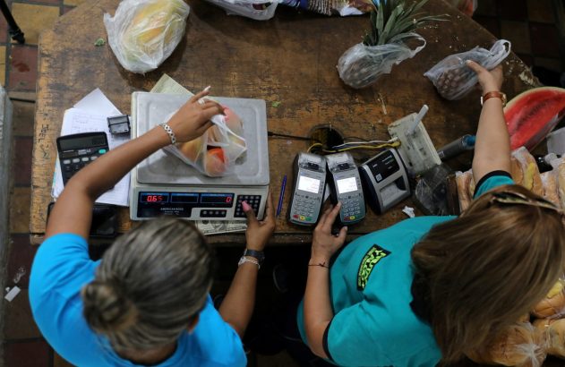 Vista aérea: duas mulheres em balcão de mercado. Uma pesa frutas na balança, a outra usa terminais de pagamento.