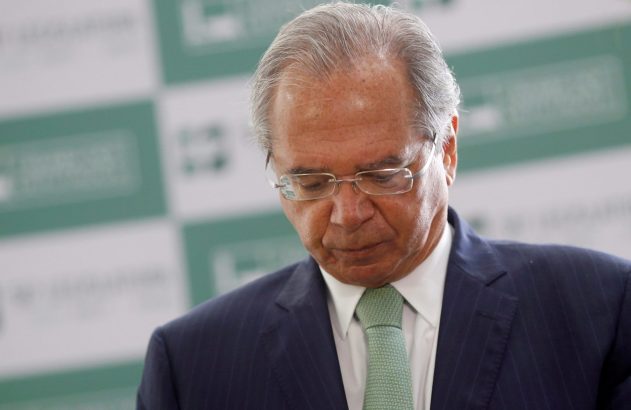 Homem de cabelos grisalhos, óculos, terno azul e gravata verde, olhando para baixo. Fundo desfocado de evento oficial.