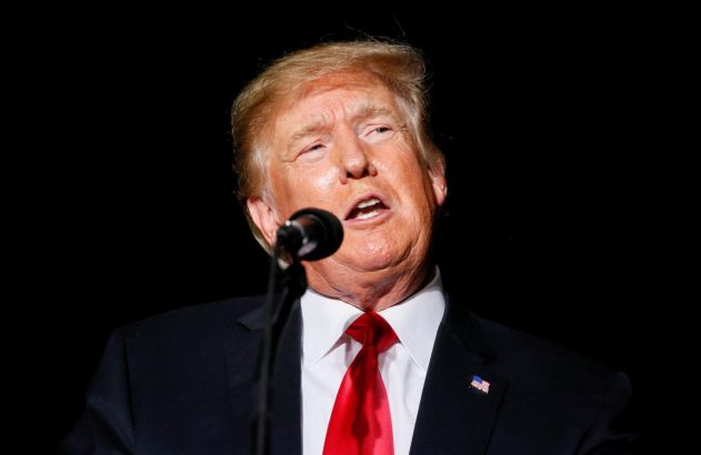 Donald Trump, de terno, gravata vermelha e broche da bandeira americana, falando ao microfone.