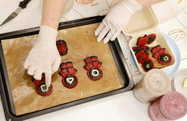Mãos enluvadas preparando biscoitos vermelhos e pretos com símbolos (círculo, triângulo, quadrado) em uma assadeira.