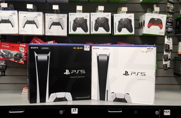 Duas caixas de PS5 (digital preta $399.99, padrão branca $499.99) em loja de games, com controles acima.