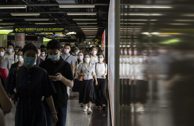 Multidão de pessoas com máscaras faciais caminha em passagem subterrânea movimentada. Placa de saída visível.