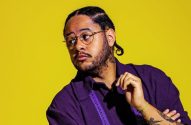 Homem de óculos, camisa roxa e tranças, olhando para a esquerda com a mão no rosto, em fundo amarelo.