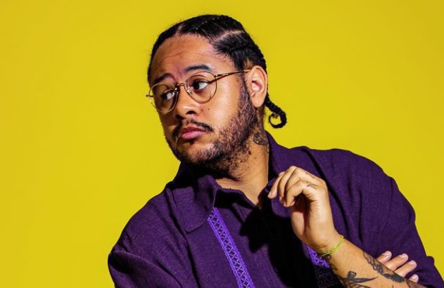 Homem de óculos, camisa roxa e tranças, olhando para a esquerda com a mão no rosto, em fundo amarelo.