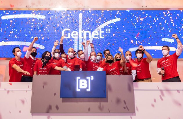 Equipe Getnet, em camisas vermelhas, celebra com confetes o IPO na Bolsa B3, sob o logo da empresa no telão.