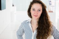 Mulher jovem sorrindo, com cabelo castanho encaracolado e camisa listrada azul e branca.