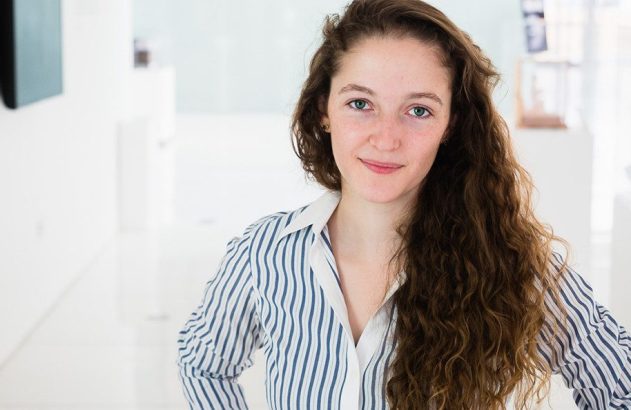 Mulher jovem sorrindo, com cabelo castanho encaracolado e camisa listrada azul e branca.