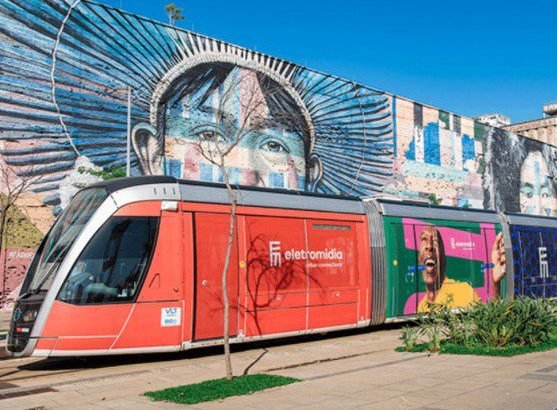 Um VLT moderno vermelho e colorido com anúncios em frente a um grande mural de arte urbana com um rosto em destaque.