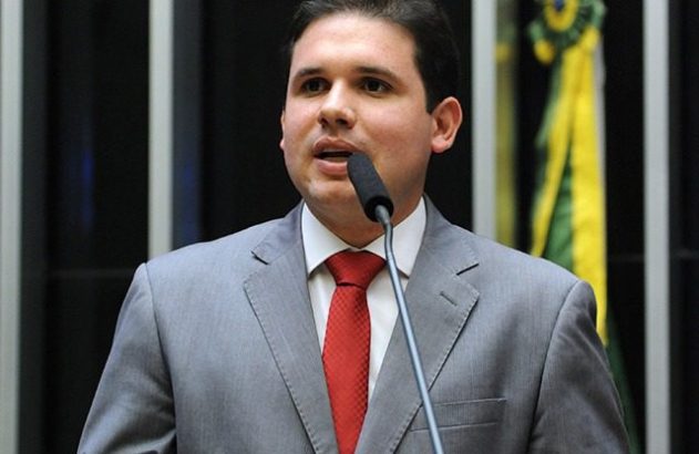 Um homem de terno cinza e gravata vermelha falando em microfone. Ao fundo, detalhes de ambiente formal e bandeira do Brasil.