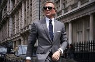 Daniel Craig como James Bond, de terno cinza e óculos escuros, andando em rua de Londres com prédios e carros.