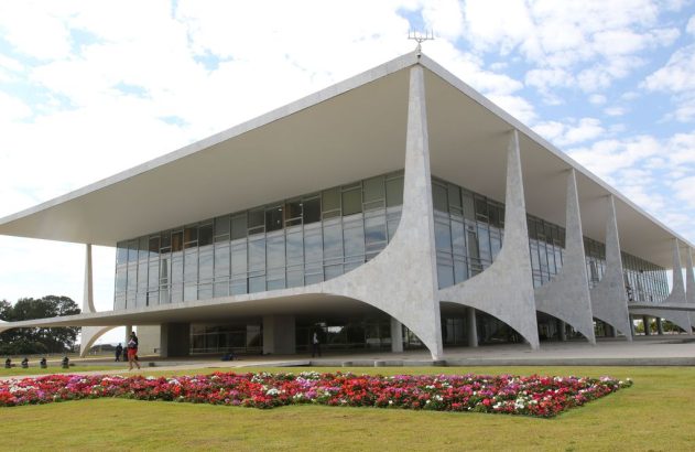Edifício moderno branco com fachada de vidro e colunas curvas, gramado verde com flores e pessoas ao redor.