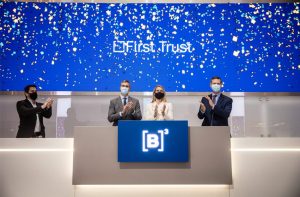 Quatro pessoas de máscara aplaudem em evento. No telão, 'First Trust' e confetes. No pódio, logo '[B]³'.