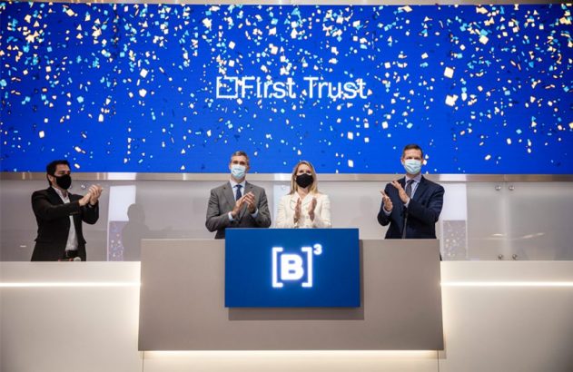 Quatro pessoas de máscara aplaudem em evento. No telão, 'First Trust' e confetes. No pódio, logo '[B]³'.