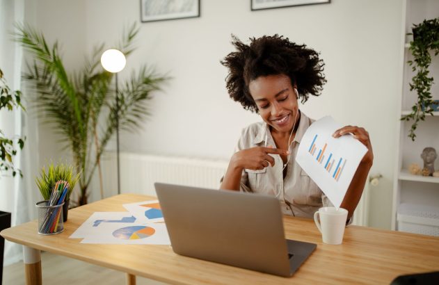 Mulher sorrindo com fones, mostra gráfico em papel para laptop, simulando chamada de vídeo.