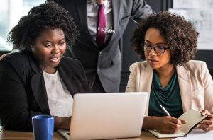 Duas mulheres e um homem em reunião. Elas analisam um laptop, enquanto uma faz anotações.