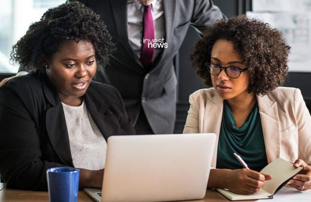 Duas mulheres e um homem em reunião. Elas analisam um laptop, enquanto uma faz anotações.