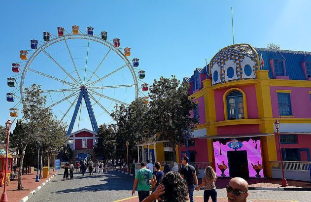 Roda-gigante e edifício colorido em um parque de diversões. Pessoas caminham em dia ensolarado.