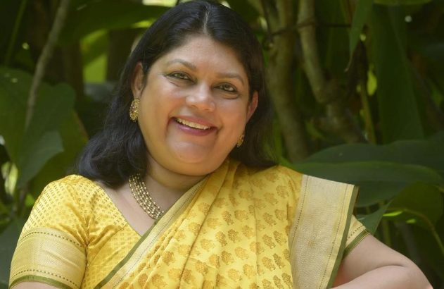 Mulher sorrindo em um saree amarelo com fundo verde de folhagens.
