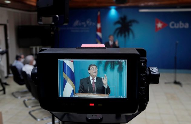 Monitor de vídeo exibe homem discursando em púlpito, com bandeira e logo de Cuba ao fundo.