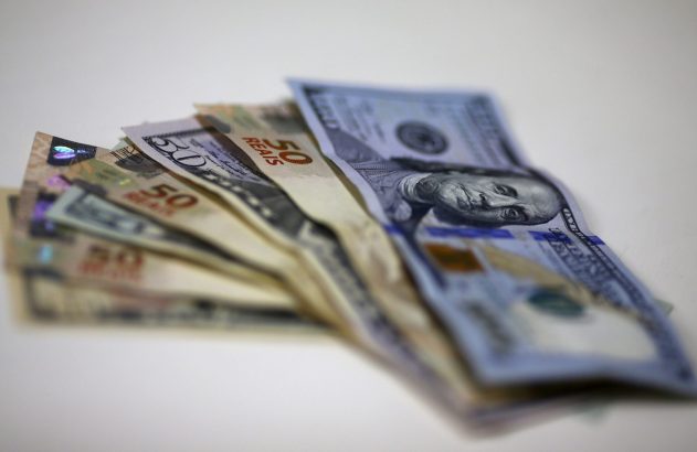 Cédulas de 100 dólares americanos e 50 reais brasileiros, dispostas em leque, sobre fundo branco.