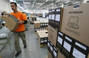 Homem move caixa de notebook em fábrica. Pilhas de caixas e esteiras em um ambiente industrial.