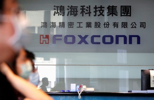 Placa de vidro com o logotipo da Foxconn e texto em chinês. Uma pessoa desfocada observa à frente.