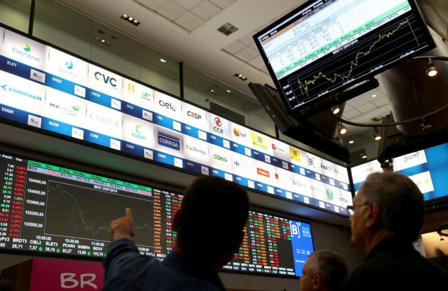 No pregão, homens olham telas da bolsa com gráficos de ações e logos de empresas, um deles apontando.