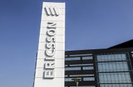 Totem branco da Ericsson com logo e nome da marca, e edifício da empresa sob céu azul.