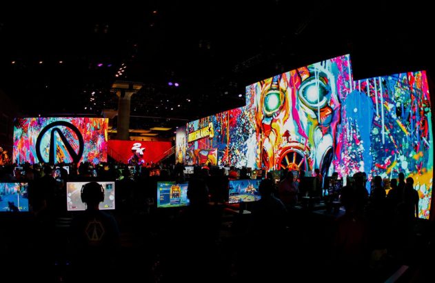 Pessoas jogam em PCs em um evento de games, com grandes telas coloridas de Borderlands 3 e 2K ao fundo.