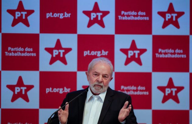 Homem de terno e barba gesticula em frente a painel xadrez do Partido dos Trabalhadores (PT).