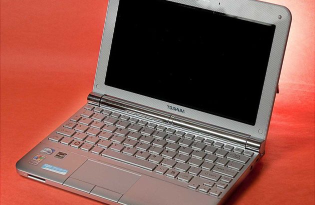 Netbook Toshiba NB205 prateado, com tela preta e teclado cinza, em um fundo vermelho.