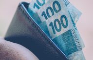 Close-up de mão segurando carteira escura com várias notas de 100 reais.