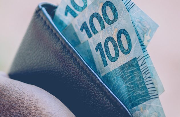 Close-up de mão segurando carteira escura com várias notas de 100 reais.