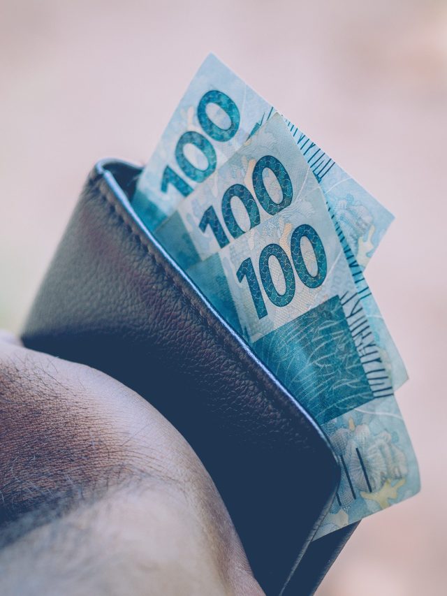 Close-up de mão segurando carteira escura com várias notas de 100 reais.