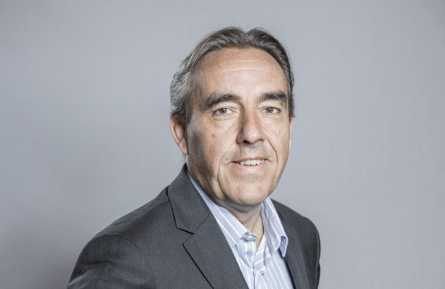 Homem sorrindo para a câmera, com cabelo grisalho e blazer cinza sobre camisa listrada, em fundo cinza.