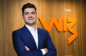 Homem de terno sorrindo e de braços cruzados, em frente ao logotipo WIZ laranja.