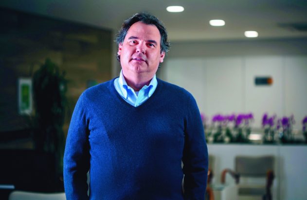 Homem de cabelos escuros e suéter azul escuro, olhando diretamente para a câmera. Fundo desfocado de escritório claro.