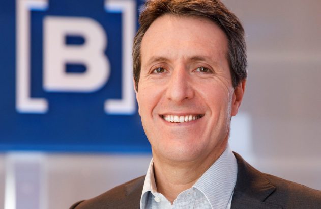 Retrato de um homem sorridente, vestindo terno e camisa, com um logo [B] branco sobre fundo azul em desfoque.