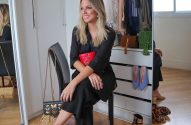 Mulher loira sorrindo, sentada em banqueta, segurando uma clutch vermelha. Bolsas e sapatos de grife adornam o espaço com espelhos.