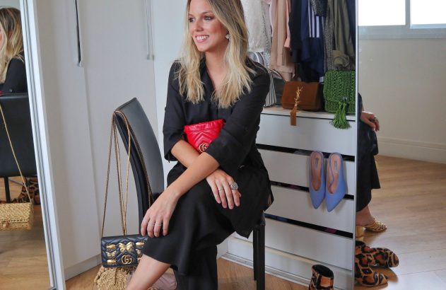 Mulher loira sorrindo, sentada em banqueta, segurando uma clutch vermelha. Bolsas e sapatos de grife adornam o espaço com espelhos.