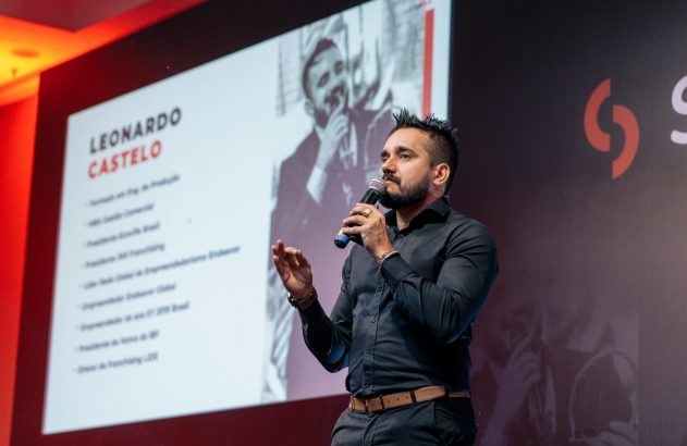 Homem de barba, camisa preta, fala em microfone no palco com tela exibindo 'LEONARDO CASTELO' e foto.