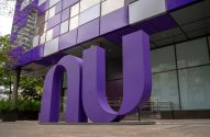 Logo gigante "NU" roxo em frente a um edifício corporativo moderno com fachada roxa e cinza.