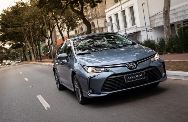 Toyota Corolla azul-acinzentado em movimento numa rua urbana. Faróis acesos, visto de frente e lateralmente.