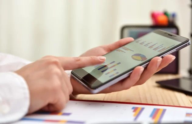 Pessoa analisando gráficos e dados estatísticos em um smartphone, com planilhas ao fundo, relacionado à análise de dados.