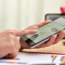 Pessoa analisando gráficos e dados estatísticos em um smartphone, com planilhas ao fundo, relacionado à análise de dados.