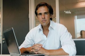 Homem de camisa branca sentado em mesa de escritório, com laptop e mãos unidas, olhando para a câmera.