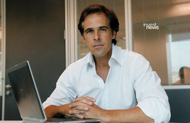 Homem de camisa branca sentado em mesa de escritório, com laptop e mãos unidas, olhando para a câmera.