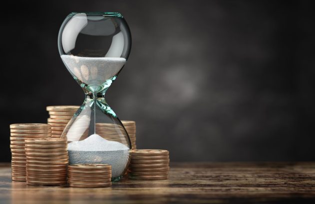 Ampulheta com areia em queda e moedas empilhadas sobre madeira.